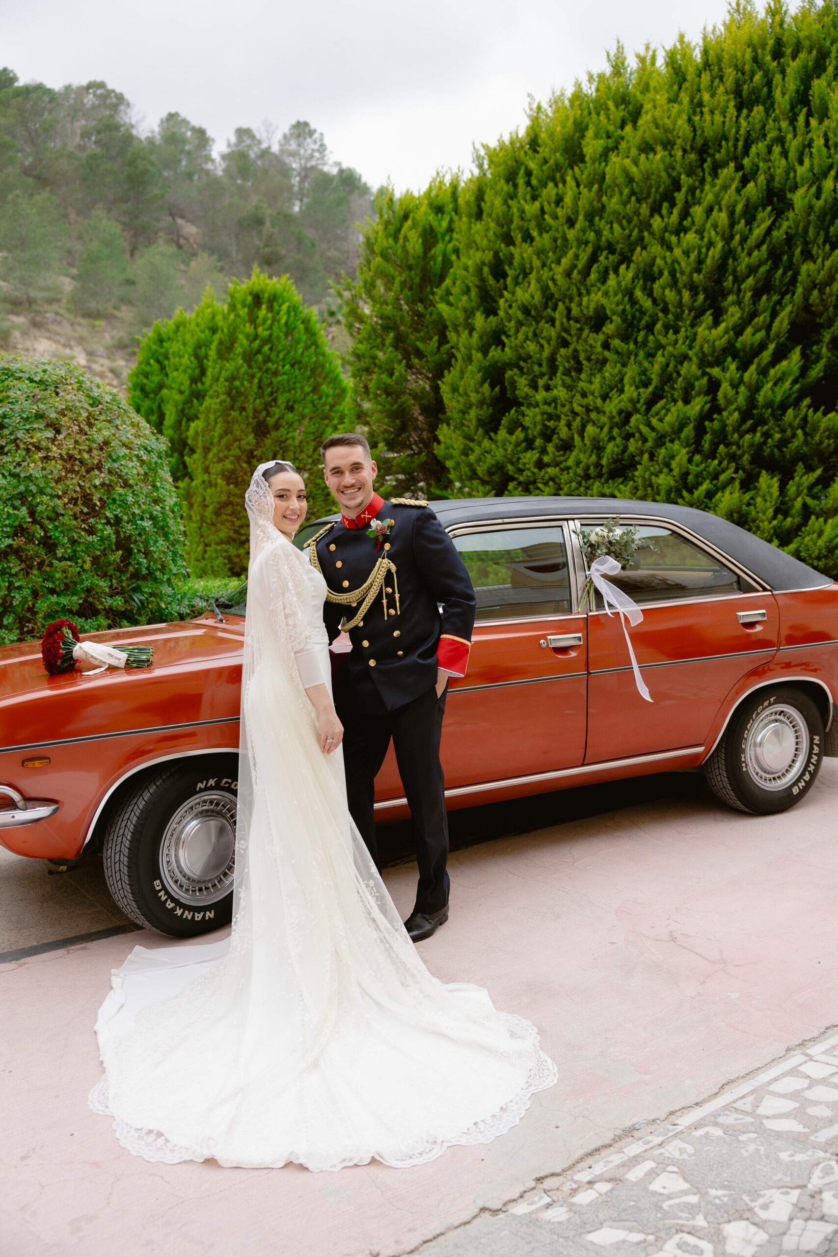 Novios junto a coche clasico rojo en boda militar en el Balneario de Archena Murcia novia con velo catedral y novio con uniforme militar